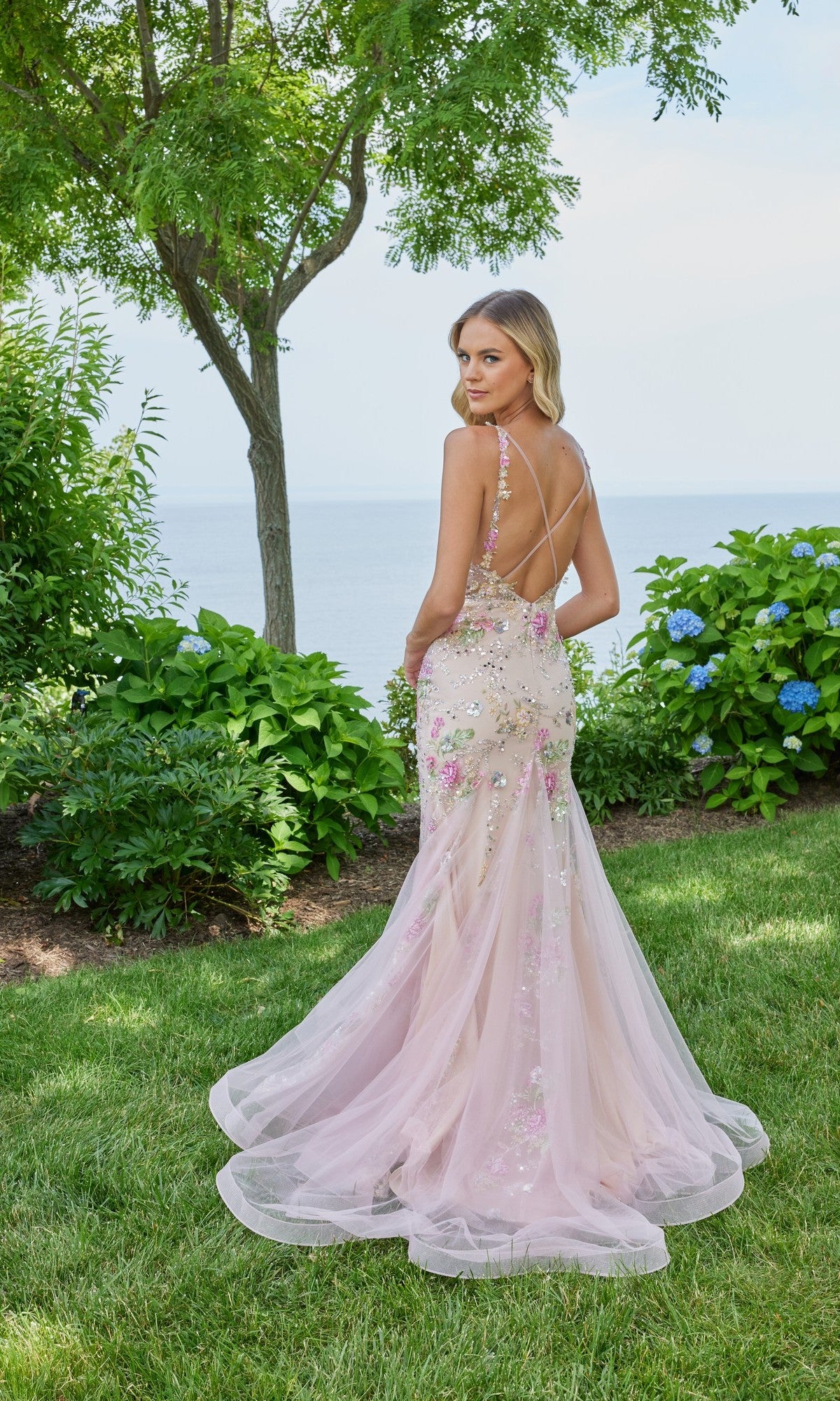 A woman in a blush pink Morilee 49130 long prom dress stands on grass by green shrubs and blue flowers, with a tree and water in the background.